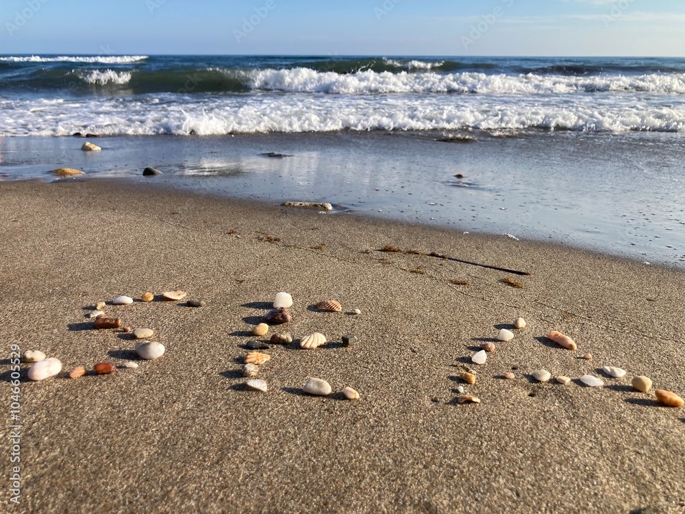 A view of a sandy beach with small seashells spelling the word “SEA” in the foreground, and gentle waves crashing in the background under a clear sky. A view of a sandy beach with small seashells spelling the word “SEA” in the foreground, and gentle waves crashing in the background under a clear sky.