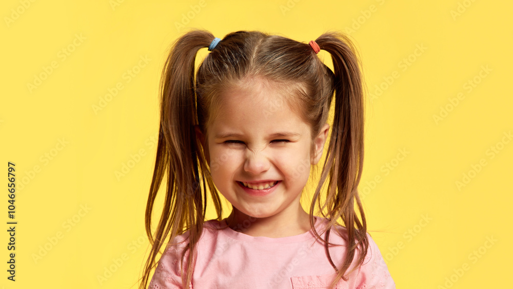 Little girl with funny pig-tails scrunches her face in exaggerated happy grin against sunny yellow background. Concept of facial-expressions, positive and negative emotions. Ad Little girl with funny pig-tails scrunches her face in exaggerated happy grin against sunny yellow background. Concept of facial-expressions, positive and negative emotions. Ad