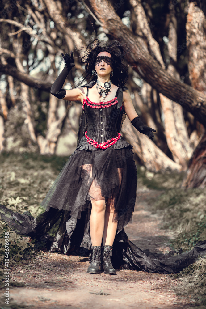 Naklejka premium A young woman in the image of a witch and sorceress, standing in a dark forest, wearing a black dress with a train and a socket on her head.