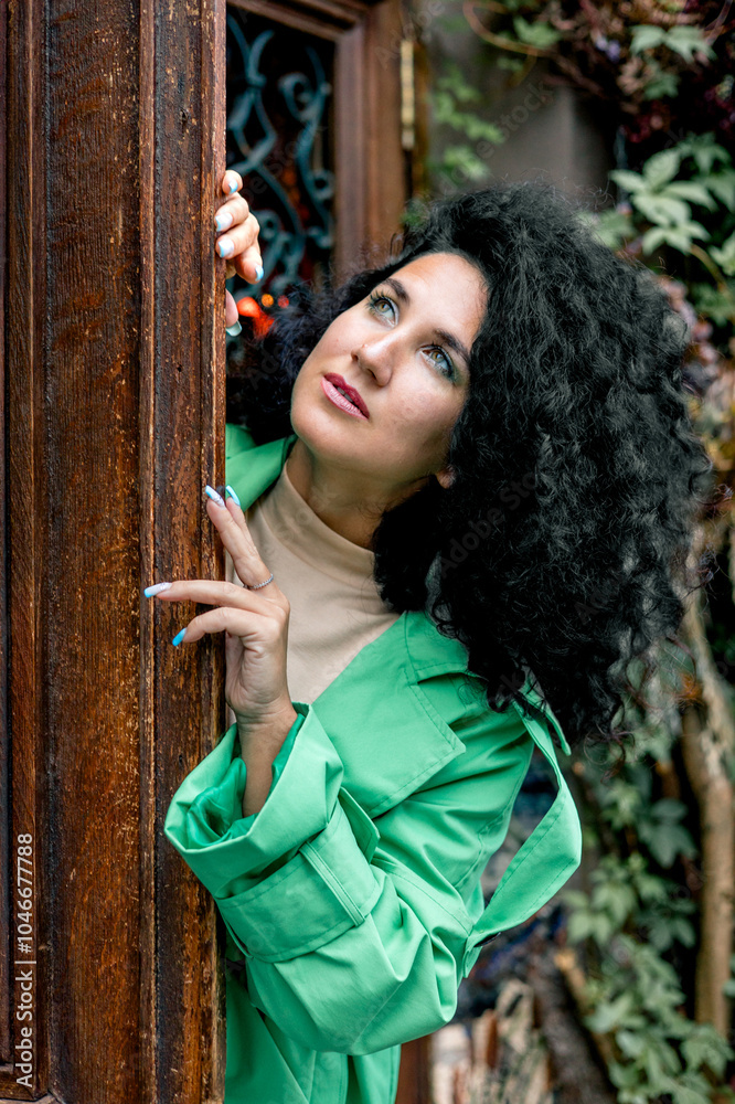Fototapeta premium Beautiful stylish young woman with curly dark hair posing in a green trench coat in an urban context. Street fashion and style.