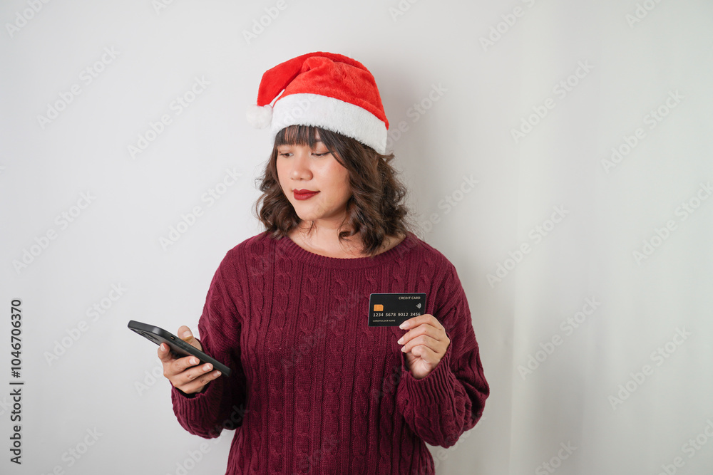 Excited young asian woman wearing santa clause hat and red long sleeved sweater is holding smartphone and credit card, isolated over white background. Concept for Christmas Holiday and New Year Party Excited young asian woman wearing santa clause hat and red long sleeved sweater is holding smartphone and credit card, isolated over white background. Concept for Christmas Holiday and New Year Party