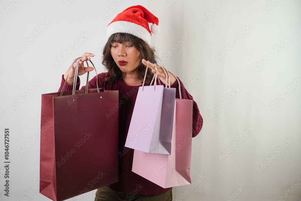 Excited young asian woman wearing santa clause hat and red long sleeved sweater is carrying many shopping bag, isolated over white background. Concept for Christmas Holiday and New Year Party. Excited young asian woman wearing santa clause hat and red long sleeved sweater is carrying many shopping bag, isolated over white background. Concept for Christmas Holiday and New Year Party.