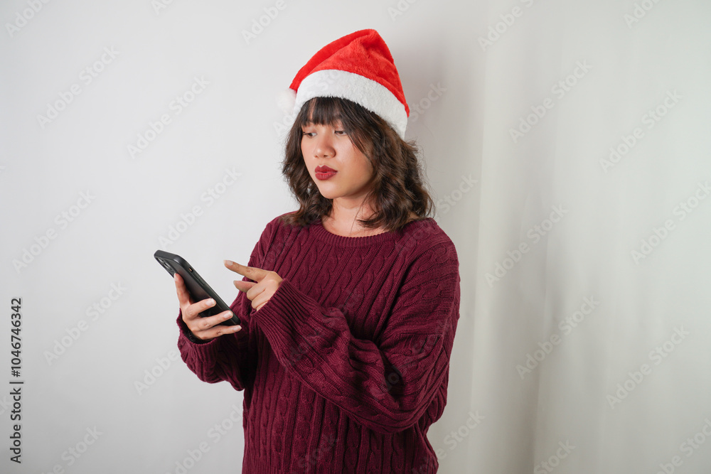 Excited young asian woman wearing santa clause hat and red long sleeved sweater is using smartphone, isolated over white background. Concept for Christmas Holiday and New Year Party Excited young asian woman wearing santa clause hat and red long sleeved sweater is using smartphone, isolated over white background. Concept for Christmas Holiday and New Year Party