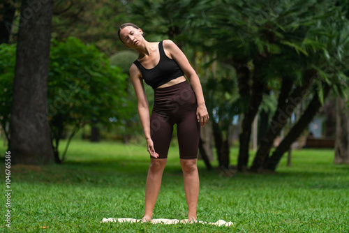 Young woman performs neck and body exercises on a mat in the park, smart fitness. Promote health and well-being. Girl in full-length sportswear performs a workout in the park. photo