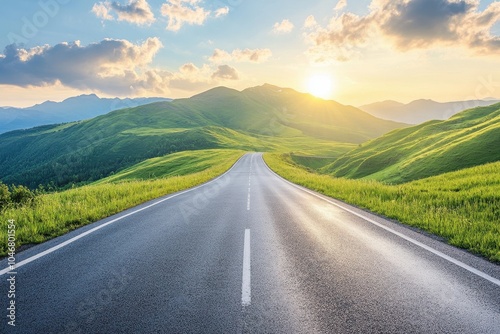 A winding asphalt road leads through a lush green valley towards a distant sunset with mountains in the background.