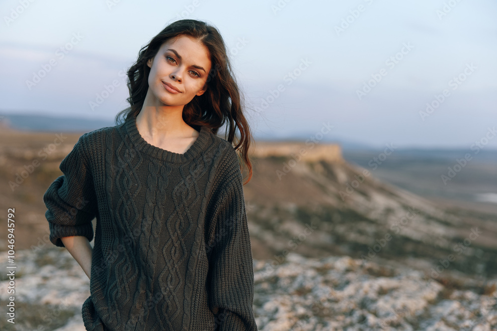 Serene woman in cozy sweater stands on hill, gazes at camera with majestic mountain backdrop Serene woman in cozy sweater stands on hill, gazes at camera with majestic mountain backdrop
