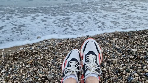 Women's legs in sneakers on the background of a pebble beach with sea waves.