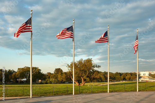 A row of four American flags stand tall, waving gracefully in the evening breeze against a backdrop of a partly cloudy sky. 