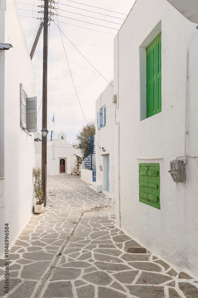 Fototapeta premium Serene alley in Paros, Greece, with green shutters, white-washed houses, and traditional Cycladic charm, bathed in natural light under a clear sky.