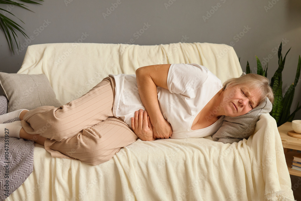 Senior woman lying on couch at home experiencing painful abdominal discomfort due to gastrointestinal cramps or nausea enduring stomach pain and inflammation feeling ill and distressed Senior woman lying on couch at home experiencing painful abdominal discomfort due to gastrointestinal cramps or nausea enduring stomach pain and inflammation feeling ill and distressed