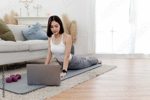 Asian woman stretching body on yoga mat in living room. Happy female working out while watch training tutorial vdo on laptop computer at home. Sporty girl doing exercise. Healthy and sport hobby photo