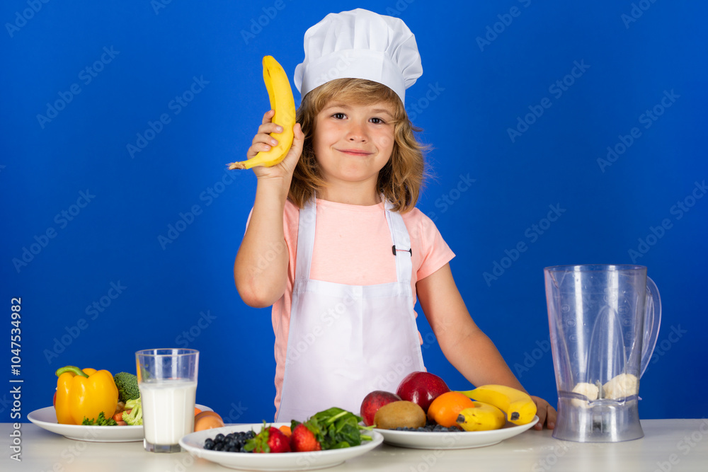 Child wearing cooker uniform and chef hat preparing vegetables hold banana on kitchen, studio portrait. Cooking, culinary and kids food concept. Child wearing cooker uniform and chef hat preparing vegetables hold banana on kitchen, studio portrait. Cooking, culinary and kids food concept.