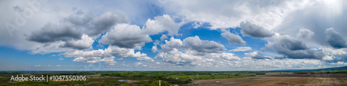 Aerial panorama of a flat prairie scene under a blue sky full of white clouds.
