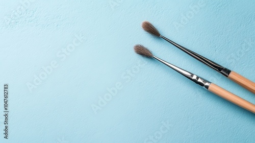 A pair of fluffy paintbrushes with wooden handles lie on a textured light blue background, offering an artistic vibe suitable for creative enthusiasts.