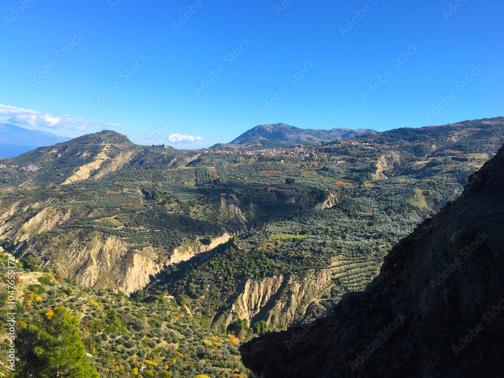 Obraz premium Aerial view of the foothills from the top of the mountain. Old olive groves, green fields, road bends among the slopes, mountain peaks under the blue sky. Greece, Peloponnese.