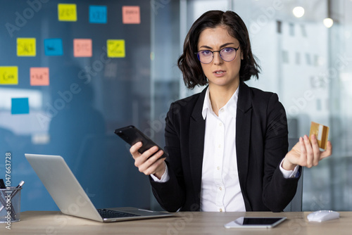 Businesswoman looking at phone and credit card with concern, considering online transaction. Office setting with laptop, highlighting concept of finance, security, and technology. photo