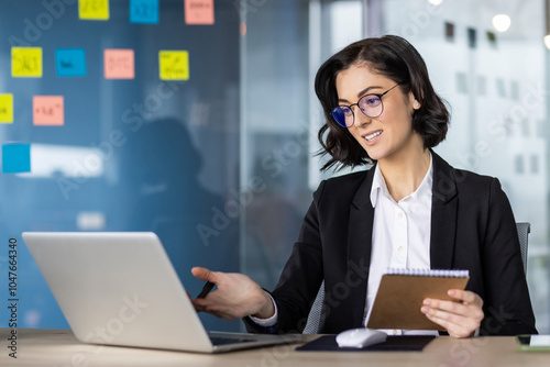 Businesswoman engaged in online meeting using laptop, holding notebook. Office setting with glass wall and sticky notes. Professional attire, conveying leadership, communication, technology.