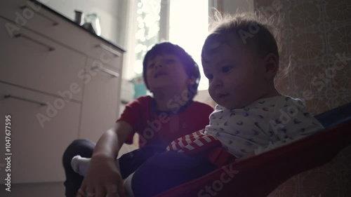 Big brother interacting with baby in a bouncer, smiling at each other, joyful sibling bonding moment, playful family atmosphere in the kitchen photo