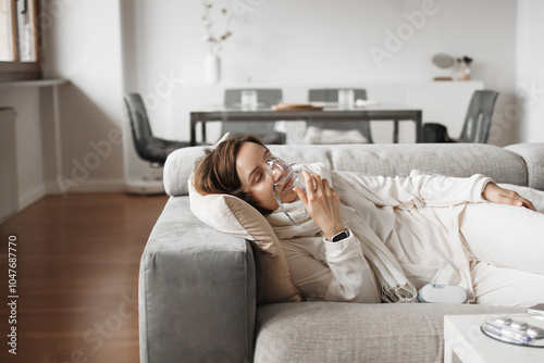 Serene Woman Breathing Through Oxygen Mask And Using Oxygen Concentrator For Respiratory Therapy At Home, Finding Comfort And Relief On Her Living Room Sofa