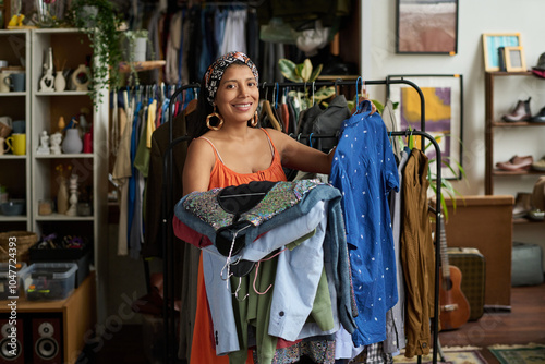 Young cheerful female owner of thrift shop or customer holding stack of casual attire from new collection of secondhand clothes