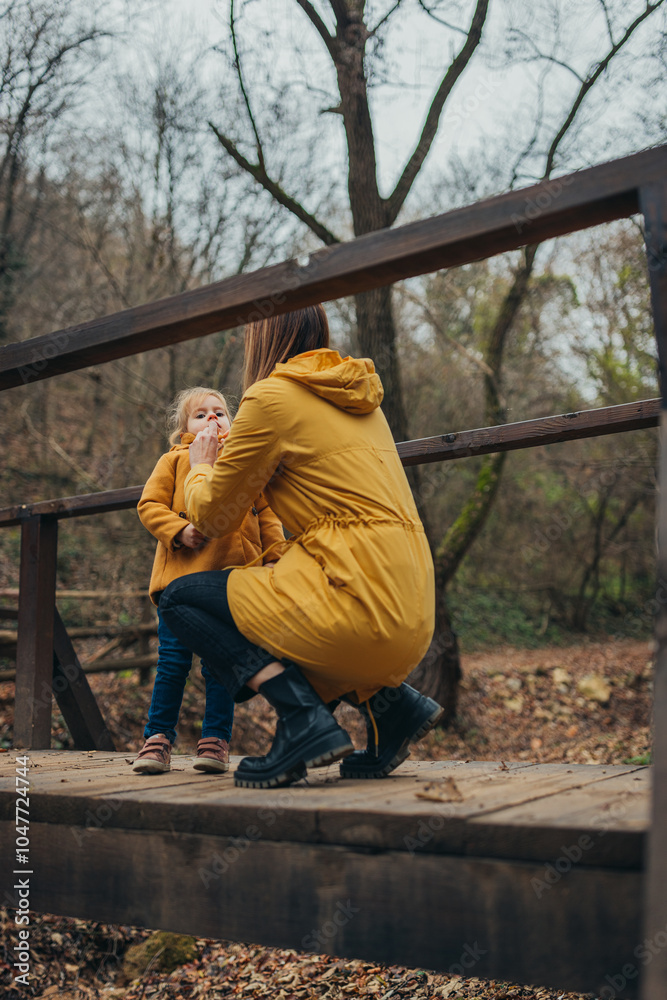A woman assists her child on a wooden bridge in a tranquil forest, both wearing yellow coats, highlighting a moment of care and warmth within nature's embrace. A woman assists her child on a wooden bridge in a tranquil forest, both wearing yellow coats, highlighting a moment of care and warmth within nature's embrace.