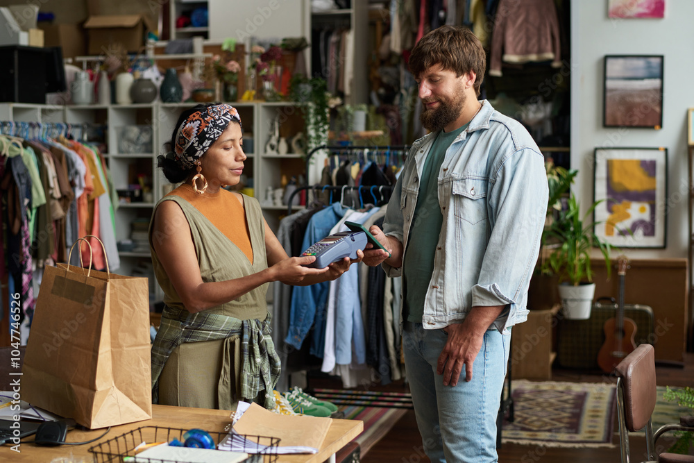 Young bearded male consumer in casualwear holding smartphone over payment terminal held by female shop assistant Young bearded male consumer in casualwear holding smartphone over payment terminal held by female shop assistant