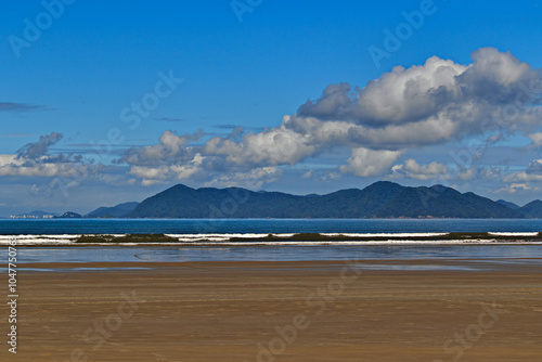 wide strip of sand during low tide, in the background a calm sea and cumulus clouds over the mountains of Serra do Mar