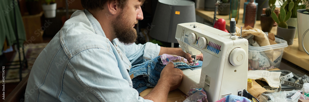 Banner of young man sitting by workplace with electric sewing machine and stitching textile pocket to used jeans or denim jacket