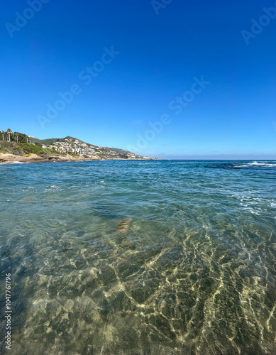 A breathtaking view of crystal-clear ocean water at Laguna Beach, with gently rolling waves and a cloudless blue sky. The sandy seafloor is visible through the transparent water.