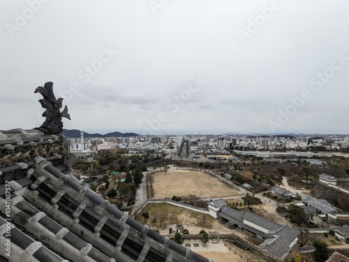 Himeji, Japan - 12.19.2023: A fish sculpture, or the shachi, on the rooftop of the Main Keep in Himeji Castle, overlooking the castle complex, gardens, entrance, and modern Himeji city under clouds