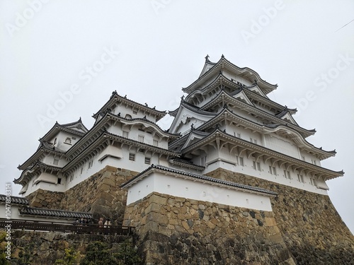 Himeji, Japan - 12.19.2023: Facade of the Main Keep on the heightened stone wall in Himeji Castle with people waiting to enter under a cloudy sky