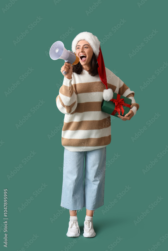 Happy young woman in Santa hat with Christmas gift shouting into megaphone on green background Happy young woman in Santa hat with Christmas gift shouting into megaphone on green background