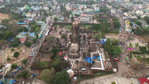 Aerial daytime view of Lingaraj Temple in Odisha, with main spire, smaller shrines, lush greenery, and bustling festival streets, contrasting nearby colorful buildings. photo