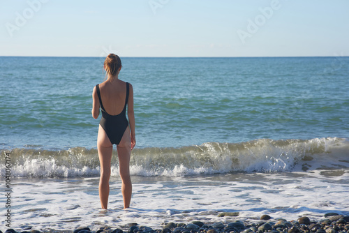 A woman in a stylish black swimsuit stands by the shoreline, gazing at the horizon on a sunny day. Waves gently crash at her feet, capturing a moment of relaxation and connection with nature.