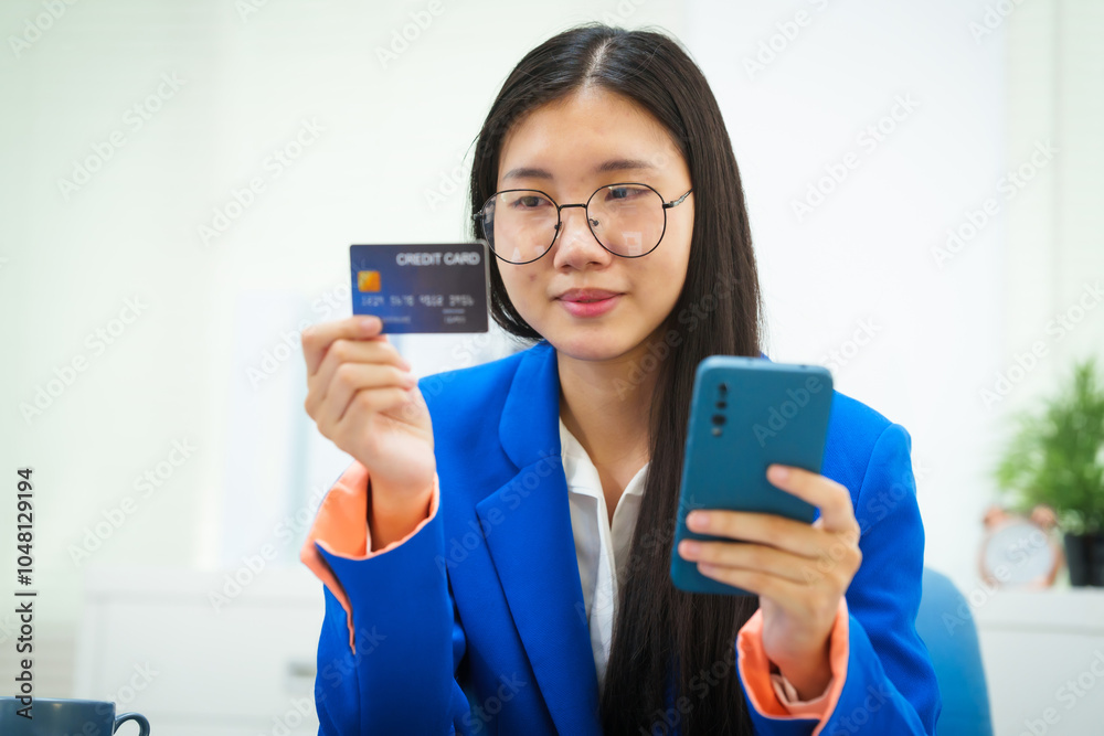 A young businesswoman sits at her office desk, smiling as she holds a credit card while shopping online. Her cheerful demeanor reflects her satisfaction with managing her work and purchases