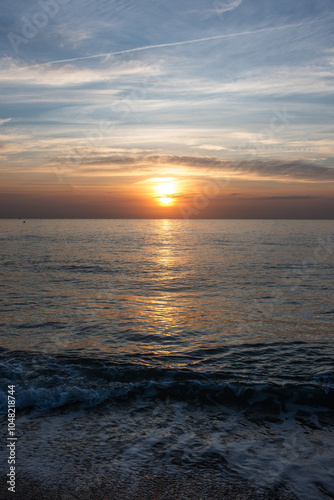 Vertical shot of a sunset at sea with the silhouette of a lighthouse on the horizon, silver and orange colored