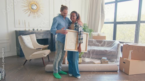 Young diverse lovers African American woman and Caucasian man are relocating or moving to new home and holding painting to update interior are stands in living room near boxes with personal belongings