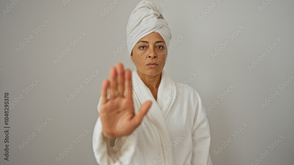 Middle age woman in a white bathrobe and towel on head standing against a white background with her hand raised in a stop gesture Middle age woman in a white bathrobe and towel on head standing against a white background with her hand raised in a stop gesture