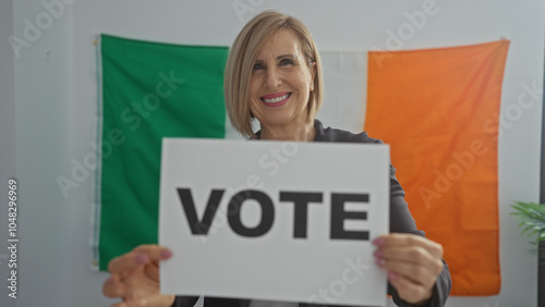 Photography A beautiful mature woman holding a vote sign stands indoors with an irish flag in the background, encouraging electoral participation