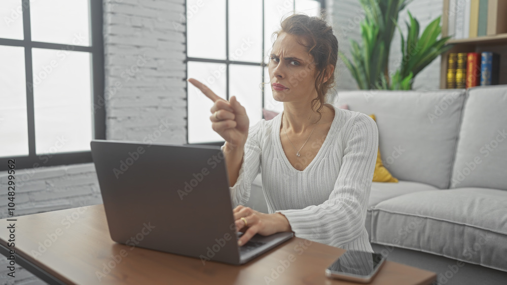 Obraz premium A concerned young woman points at a laptop screen while sitting in a modern living room, indicating an issue or question.