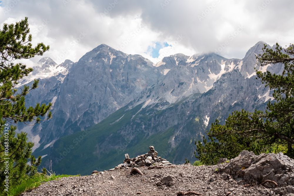 Obraz premium Breathtaking alpine landscape with trail leading to rocky outcrop. Small cairn of stones is piled at the edge offering panoramic view of mountain ridges of Albanian Alps (Accursed Mountains), Albania