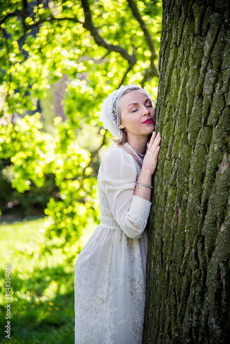 portrait of beautiful blonde smiling  woman in vintage white dress and mutch  with closed eyes hugging tree in the summer garden. Wedding day photo