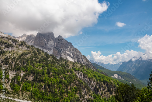 Scenic hiking trail through alpine pine forest. Majestic steep mountain peaks of massif Zhaborret shrouded in clouds, Albanian Alps (Accursed Mountains), Valbone Valley National Park, Albania. Hike photo