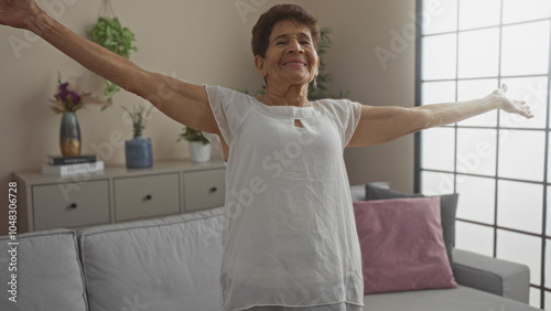 Elderly hispanic woman enjoying a moment of joy with arms outstretched in a cozy, modern living room of her home.