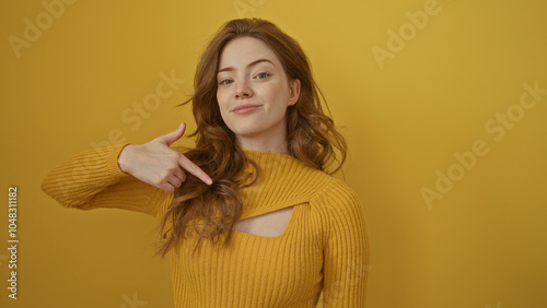 A young, attractive, blonde woman points to herself against an isolated yellow background wall, exuding confidence and beauty.