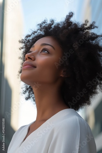 Woman with curly hair gazes upward in a cityscape under a bright sky during t...
