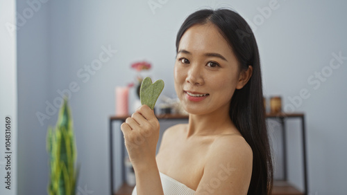 Asian woman holding a jade gua sha stone in a serene wellness spa room, symbolizing beauty and relaxation. photo