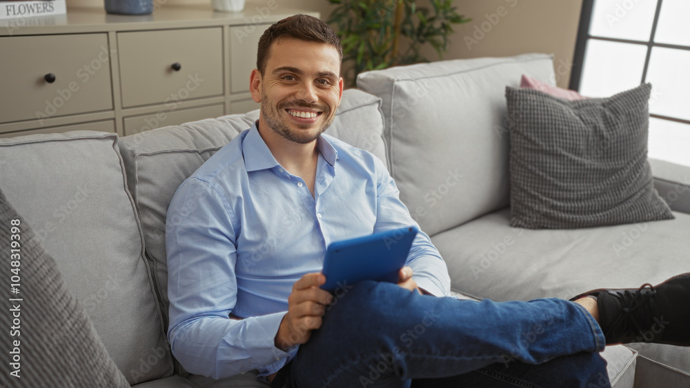 Handsome young hispanic man with a beard sitting in a living room holding a tablet and smiling. Handsome young hispanic man with a beard sitting in a living room holding a tablet and smiling.