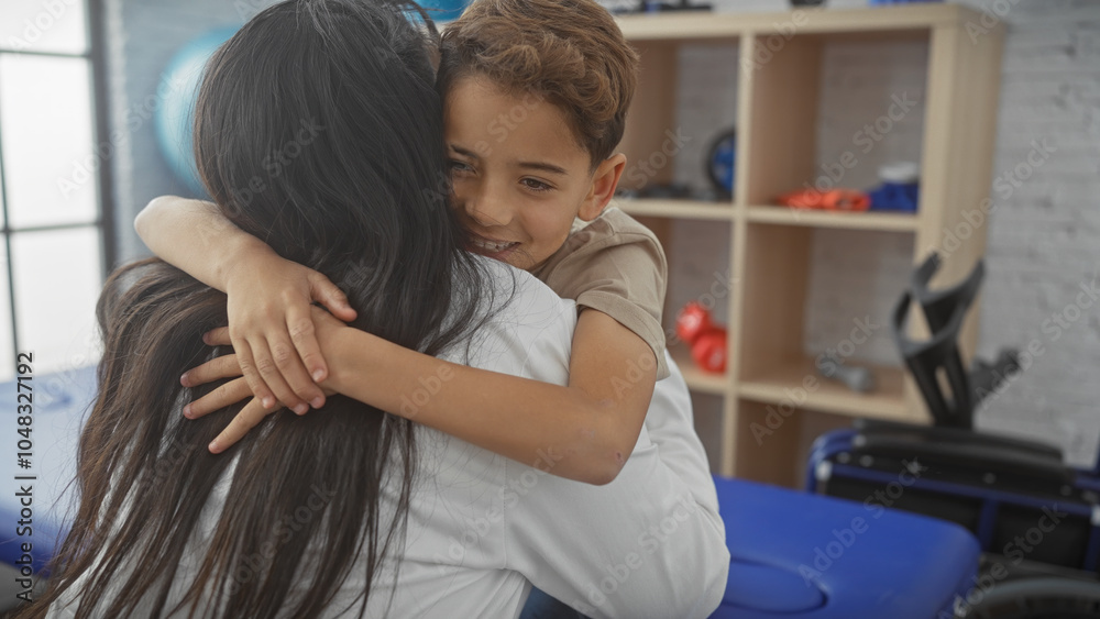 In a rehab clinic, a caring woman embraces a smiling boy, symbolizing recovery and support in a medical indoor setting. In a rehab clinic, a caring woman embraces a smiling boy, symbolizing recovery and support in a medical indoor setting.
