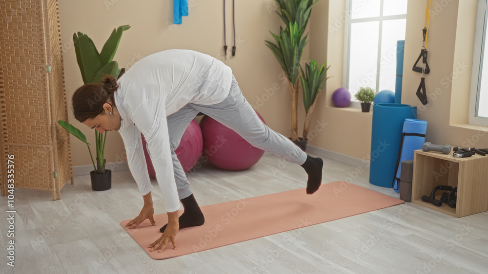 Young man stretching in a gym with exercise equipment, performing a yoga position on a mat surrounded by workout accessories and plants. Young man stretching in a gym with exercise equipment, performing a yoga position on a mat surrounded by workout accessories and plants.